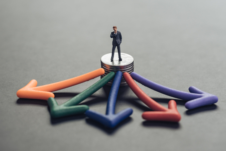 Financial investment choices, consultant for business decision concept, miniature people businessman standing and thinking on stack of coins with multi direction color arrow pointing out.の写真素材