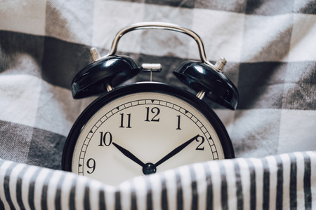Black retro alarm clock sleeping on pillow with blanket metaphor of insomnia, late at work, well sleep with time countdown or wake up in the morning.の写真素材