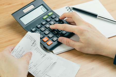 Cost and expense calculation or bill payment concept, hand put finger on calculator and black pen on paper notepad and holding pile of bills or receipt in the left hand on wooden tableの写真素材
