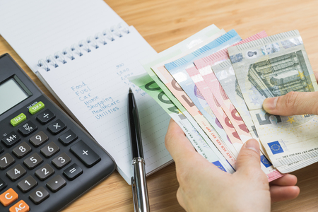Expense and budget concept, woman hand holding Euro banknote money with list of expense in small notepad and calculator on wooden table.の写真素材