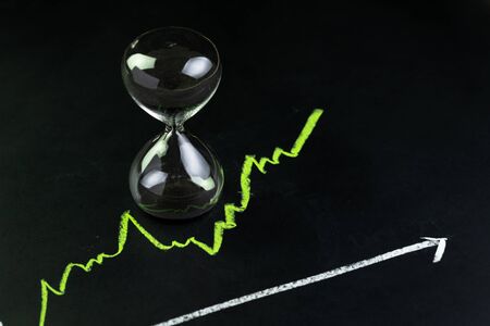Time for invest or long term investment concept, hourglass or sandglass with black sand inside on stock market green line rising chart drawing with chalk on black chalkboard.の写真素材