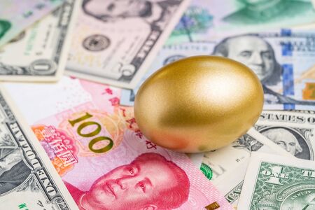 Shiny golden egg on pile of Chinese Yuan and US America dollar banknotes money metaphor of finding the good stock with high dividend or success investment in stock market from trade war concept.の写真素材