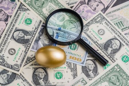 Magnifying glass with shiny golden egg under pile of US America dollar banknotes money metaphor of precious wealth, finding good stock with high dividend or success investment in stock market concept.の写真素材