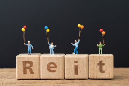 Reit, Real estate investment trust concept, happy miniature people holding balloons on cube wooden block with alphabet combine the word Reit on dark black background.の写真素材