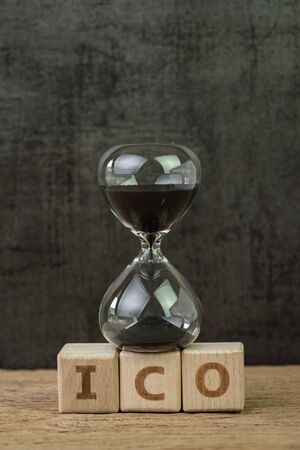 ICO, Initial Coin Offering for crypto currency to buy and sell on public concept, sandglass or hourglass on wooden cube block with alphabet building the word ICO on wood table, dark black background.の写真素材