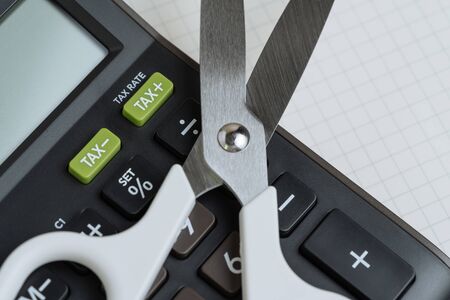 Tax cut, budget reduction, debt cutout concept, white scissors on black calculator with green button with tax minus and tax plus label on grid line paper note book.の写真素材