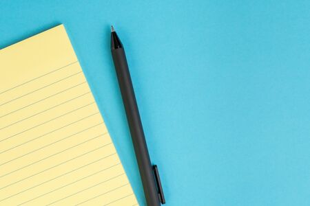 Small yellow paper notepad with blank page, pen on solid blue background using as meeting note,...