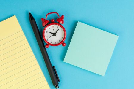 Small yellow paper notepad with blank page, alarm clock, pen and sticky note on solid blue background using as meeting note, writing message or task list on working project.の写真素材