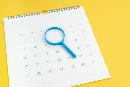 Schedule, focus date or special event day concept, blue magnifying glass on white clean calendar on solid yellow background.の写真素材