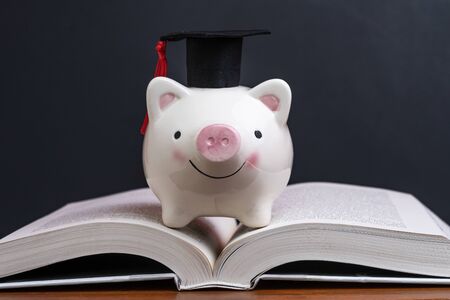 Scholarship, savings for university, cost of knowledge or financial education concept, pink smiling piggy bank with graduation hat on top of opening book with dark blackboard or chalkboard background.の写真素材