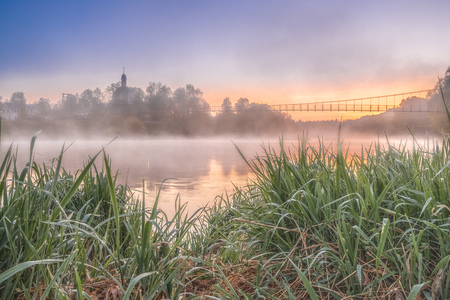 Colorful view of the church and suspension bridge from the opposite bank of the river. Dawn cool morning. Grass with drops of dew and fog over the water. Suspension bridge dissolving in fog.の写真素材