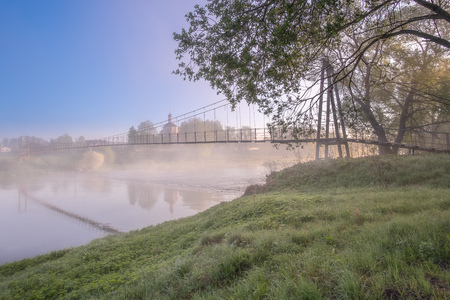 Colorful view of the church and suspension bridge from the opposite bank of the river. Dawn cool morning. Grass with drops of dew and fog over the water. Suspension bridge dissolving in fog.の写真素材