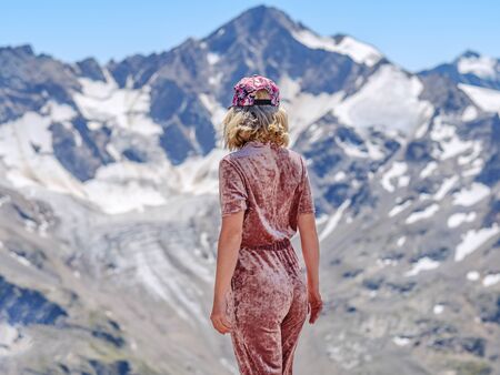 Rear view of a blonde in a baseball cap posing against a background of snowy mountain peaks of the Caucasus. Sunny summer day in the mountains.の写真素材