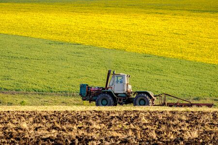 An agricultural tractor in the evening sun plows the field with a plow after harvesting wheat. In the background are fields of bright yellow sunflowers.の写真素材