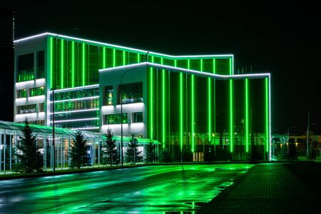 Modern tech building framed by a line of green neon lights. A bewitching view of the night city with reflection of neon lights on a wet road.の写真素材