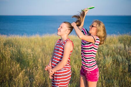 A young woman in glasses is combing the hair of a teenage girl on a sunny summer day on the lake. Mother and daughter in striped clothes. Happy family.の写真素材