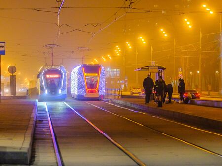Moscow.Russia. December 26, 2019. Trams in the fog lit by a network of LEDs arrive at a bus stop with passengers. Environmentally friendly public transport, decorated in honor of the New Year holidaysのeditorial素材