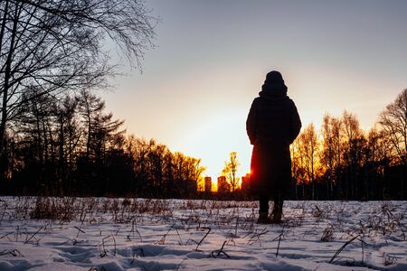 Silhouette of a female figure standing in the snow in the backlight of the setting sun in winter. A...