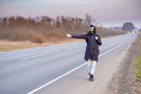 A girl in dark clothes, a gas mask and a hood on her head. Cloudy day in the country. She raises her hand and tries to stop the passing car. Escaping from a zone infected with a coronavirus.の写真素材