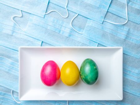 Colored Easter eggs on a white rectangular ceramic plate on a background of blue medical masks. Easter holiday concept during quarantine epidemic. Copy spaceの写真素材