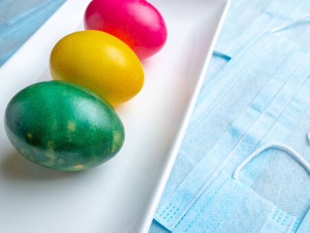 Colored Easter eggs on a white rectangular ceramic plate on a background of blue medical masks.  Easter holiday concept during quarantine epidemic. Copy spaceの写真素材