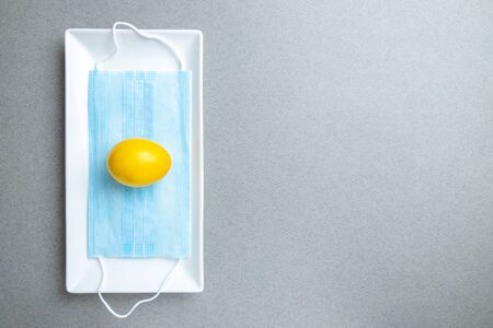 Yellow easter egg lies on a blue medical mask inside a white rectangular ceramic plate on a gray background. Flat lay. Easter holiday concept during quarantine epidemic. Copy space.の写真素材