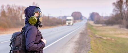 A girl in a black gas mask stands on the edge of a suburban highway. Banner. The girl is trying to stop passing cars in order to leave the city in which the epidemic of coronavirus.の写真素材