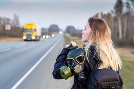 A girl with a black gas mask on his shoulder stands on the edge of a suburban highway. The girl is trying to stop passing cars in order to leave the city in which the epidemic of coronavirus.の写真素材