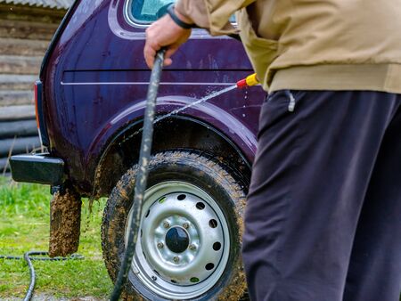 A gray-haired elderly man washes a car outdoors with a garden hose. A powerful jet of water flushes dirt from the purple body of the car, from glass, wheels and tires.の写真素材