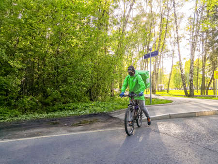 Moscow, Russia. May 15, 2020. Courier in green clothes and in a black protective mask on a bicycle in a city park on a sunny day. Food delivery in quarantined coronavirus epidemic.のeditorial素材