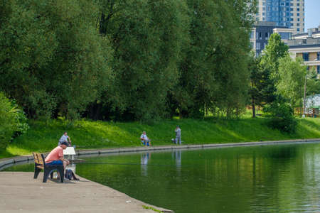 Moscow, Russia. June 12, 2020. Male fishermen are sitting on the shore of a city reservoir and fishing with fishing rods. Summer sunny day. Lush green vegetation. Active lifestyle. Outdoor hobbies.のeditorial素材