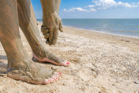 Selective focus on beautiful female foot with bright red pedicure on the beach sand. Hands smear healing mud on the ankles of the feet. Skin rejuvenation while relaxing at sea. Spa. copy spaceの写真素材