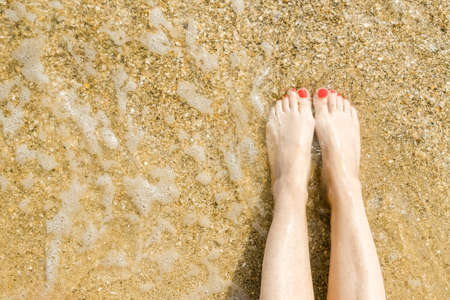 Top view of beautiful female feet with bright red pedicure on the sand of the beach. The sea wave washes womens feet. Relaxation and enjoyment during your seaside holiday. Copy space.の写真素材