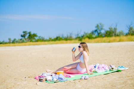 Sexy young girl in a white bathing suit sits on a mat with a mug in her hand and drinks. Girl picnic...
