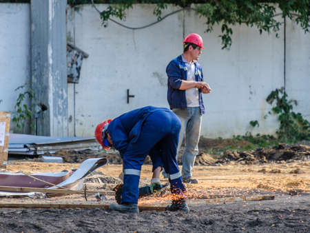 Moscow. Russia. 06 September 2020 Workers in blue uniforms and red safety helmets on a construction site are cutting metal with a grinder. Sparks of burning metal shavings fly aroundのeditorial素材