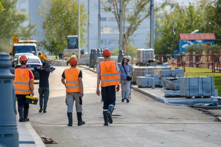 Moscow. Russia. September 06, 2020 Rear view of a group of construction workers wearing orange vests and safety helmets walking along the construction site among the building materialsのeditorial素材