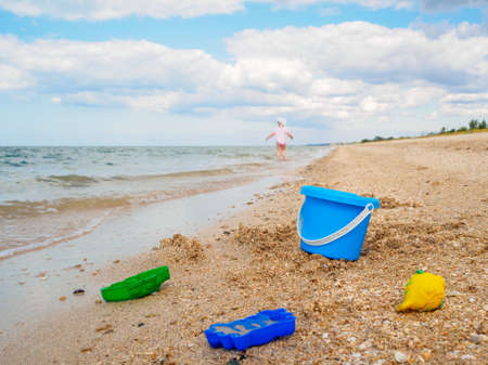 A blue plastic bucket and toys in the sand on the seashore against the background of the sky and a little girl running away into the distance. The concept of recreation with children at seaの写真素材