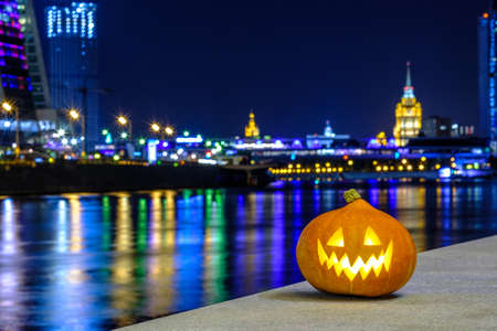 Halloween pumpkin on the background of the night city. Multi-storey buildings and skyscrapers. Blurry colored lights are reflected in the water. Night city decor with a festive Halloween theme.の写真素材