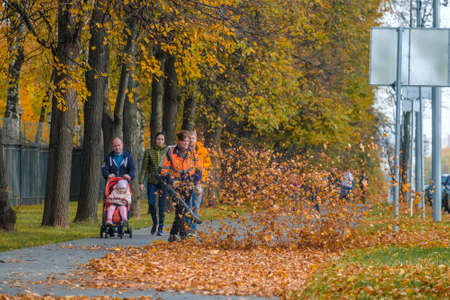 Moscow. Russia. October 11, 2020: A female utility worker uses a blower to remove fallen leaves in an alley in a park. Yellow leaves are flying in the air. Seasonal work concept.のeditorial素材
