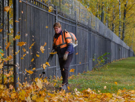 Moscow. Russia. October 11, 2020: A female utility worker uses a blower to remove fallen leaves on a lawn in a park.のeditorial素材