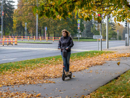 Moscow. Russia. October 11, 2020. Young woman rides an electric scooter on the sidewalk in a city park. Walk in the street on an autumn day. There are golden fallen leaves on the asphalt.のeditorial素材
