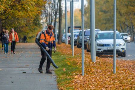 Moscow. Russia. October 11, 2020: A female utility worker uses a blower to remove fallen leaves in an alley in a park. Yellow leaves are flying in the air. Autumn sunny day. Seasonal work concept.のeditorial素材