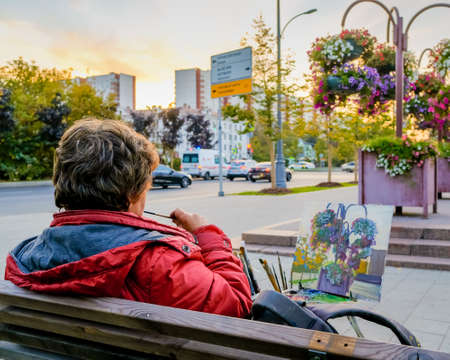 Moscow. Russia. October 1, 2020 A woman artist paints a picture with a brush and paints, sitting on a bench on a city street. Autumn evening. Flower beds with multi-colored flowers.のeditorial素材