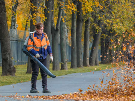 Moscow. Russia. October 11, 2020: A female utility worker uses a blower to remove fallen leaves in an alley in a park. Yellow leaves are flying in the air. Autumn sunny day. Seasonal work concept.のeditorial素材