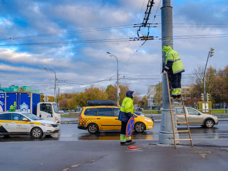 Moscow. Russia. October 19, 2020 Two workers in yellow uniforms assemble a road sign on a lamppost on a city street. A man stands on a ladder. Cloudy autumn day. Cars are driving along the road.のeditorial素材