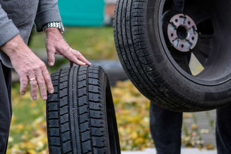 The man's hands are holding a new black rubber tire. Seasonal tire change. Autumn day. Yellow fallen leaves on the ground in the background.の写真素材