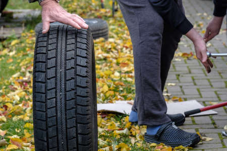 The man's hands are holding a new black rubber tire. Seasonal tire change. Autumn day. Yellow fallen leaves on the ground in the background.の写真素材