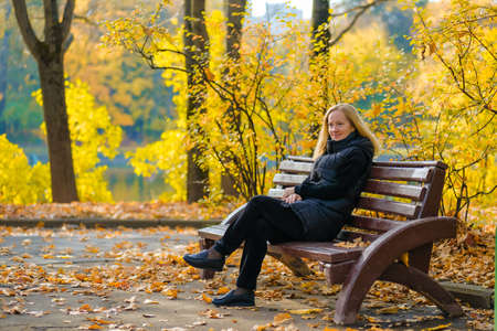 A beautiful young blonde is sitting on a bench in an autumn park.の写真素材