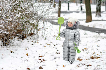 Happy child on a winter walk in the park. A funny little girl in a warm silver jumpsuit holds a toy shovel. First snowfall. Dry maple leaves peek out from under the snow. Blurred background.の写真素材