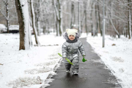 Happy child on a winter walk in the park. A funny girl in a warm silver overalls runs along the path. The baby is holding a toy shovel. First snowfall. Blurred background.の写真素材